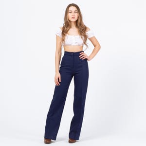 May include: A woman wearing a white off-the-shoulder crop top and high-waisted, wide-leg navy blue pants. The pants have a button closure and a tailored fit. The outfit is styled with brown shoes.