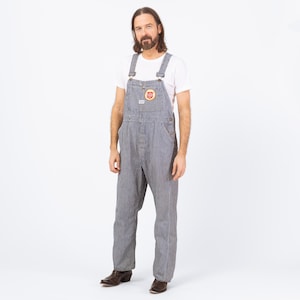 May include: Blue and white striped overalls with a white t-shirt. The overalls have adjustable straps, a front pocket with a circular patch, and button closures. The overall's inseam is approximately 30 inches.