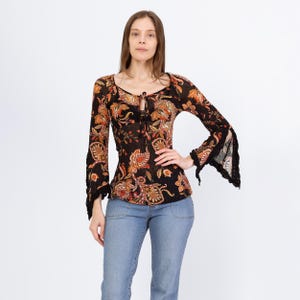 May include: A black long-sleeve top with a paisley pattern in shades of orange, red, and gold. The top features a tie-up neckline and flared sleeves with black lace trim. The model is wearing light blue jeans.