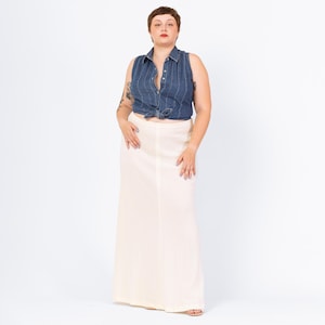 Peut inclure: Un haut sans manches en jean avec des rayures blanches verticales est noué à la taille, associé à une longue jupe crème. La tenue est portée par une personne aux cheveux bruns courts et tatouages sur le bras.