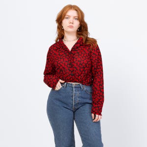 May include: A red and black button-down shirt with a repeating lip pattern. The long-sleeved shirt is paired with blue denim jeans. The model is wearing a silver necklace and earrings.