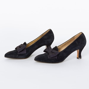 Puede incluir: Un par de zapatos de sal&oacute;n negros de ante con tacones peque&ntilde;os y lazos decorativos. Los zapatos tienen una punta afilada y un tac&oacute;n bajo. Los lazos est&aacute;n centrados en la parte delantera de los zapatos.