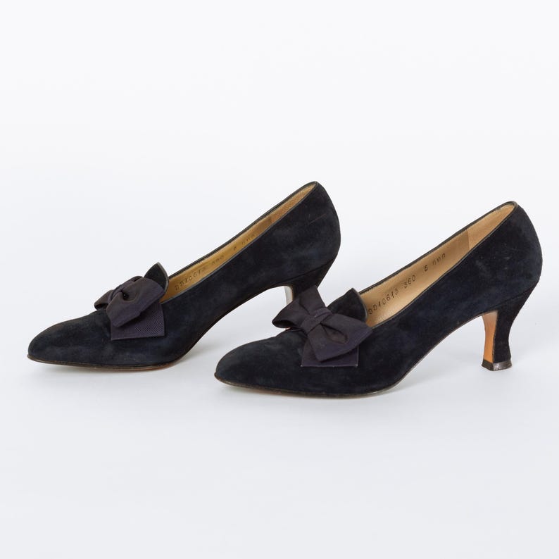 Puede incluir: Un par de zapatos de sal&oacute;n negros de ante con tacones peque&ntilde;os y lazos decorativos. Los zapatos tienen una punta afilada y un tac&oacute;n bajo. Los lazos est&aacute;n centrados en la parte delantera de los zapatos.