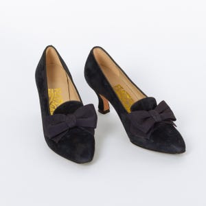 Puede incluir: Un par de zapatos de sal&oacute;n negros de ante con un lazo en el empeine. Los zapatos tienen una punta afilada y un tac&oacute;n peque&ntilde;o. El interior del zapato tiene texto dorado. Los zapatos est&aacute;n sobre un fondo blanco.