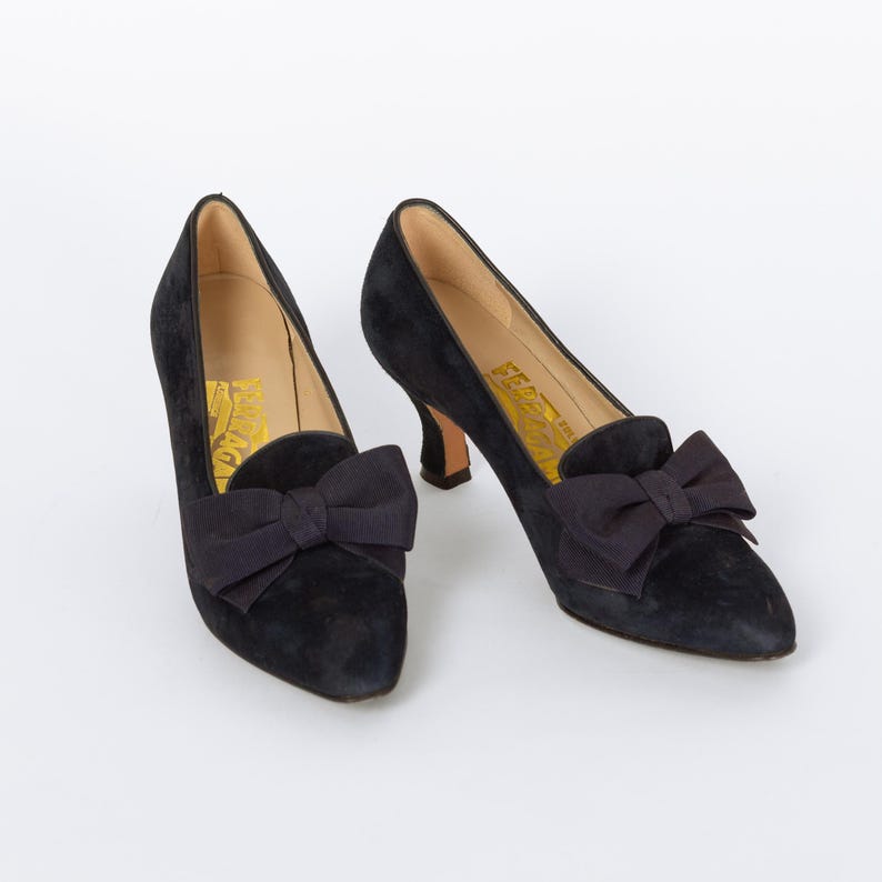 Puede incluir: Un par de zapatos de sal&oacute;n negros de ante con un lazo en el empeine. Los zapatos tienen una punta afilada y un tac&oacute;n peque&ntilde;o. El interior del zapato tiene texto dorado. Los zapatos est&aacute;n sobre un fondo blanco.