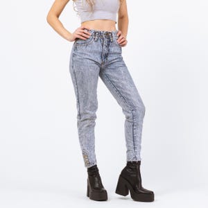 May include: Light blue acid wash jeans with a high waist and a tapered leg. The jeans have a button and zipper closure and are worn with black platform boots. The jeans have a vintage style.