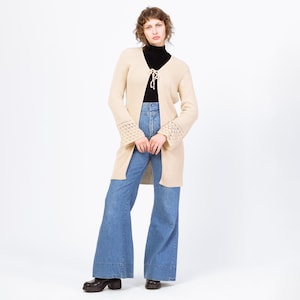 May include: A long, beige cardigan with a tie closure and crochet detailing on the sleeves. The cardigan is worn over a black turtleneck and blue flared jeans. The model is wearing black platform shoes.