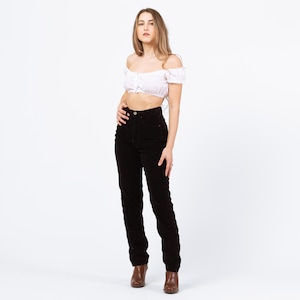 May include: A woman wearing a white off-the-shoulder crop top and high-waisted, dark brown corduroy pants. The pants have a classic, straight-leg fit. The woman is also wearing brown ankle boots.