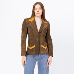 May include: A brown suede blazer with a mustard yellow collar and pocket accents. The blazer has a single-button closure and two front pockets. The model is wearing blue jeans. This vintage jacket is a stylish addition to any wardrobe.