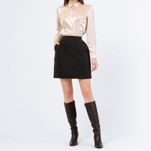 May include: A black quilted mini skirt with a satin button-down shirt. The skirt has a high waist and a straight silhouette. The shirt is a light beige color and has a classic collar.