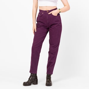 May include: A person wearing high-waisted, dark purple jeans with a button closure and pockets. The jeans are paired with black boots. The person is also wearing a white crop top.