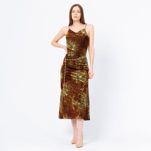 May include: A midi-length slip dress with a floral pattern in shades of brown, gold, and black. The dress features spaghetti straps and a gold chain belt. The model is wearing nude-colored heels.