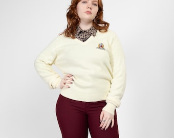 May include: Cream V-neck sweater with an embroidered emblem, worn with a patterned collared shirt and burgundy trousers. The outfit is set against a plain white background. The sweater is a classic style.
