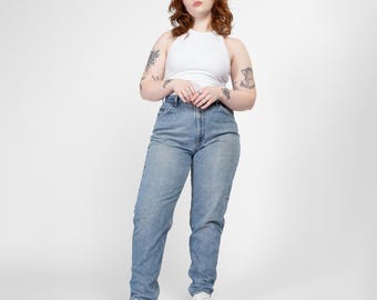 May include: A woman wearing a white tank top and light blue denim jeans. She is standing with her hands clasped in front of her.