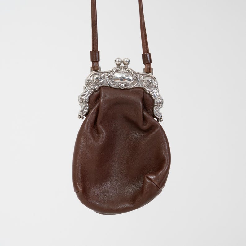 Puede incluir: Un peque&ntilde;o bolso de cuero marr&oacute;n con un marco de metal plateado que presenta un dise&ntilde;o ornamentado y repujado. El bolso tiene un cierre de boquilla en la parte superior e incluye una fina correa de cuero marr&oacute;n para llevar al hombro.