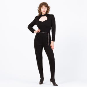 Peut inclure: Combinaison noire à manches longues avec décolleté en trou de serrure. La combinaison est ornée d'une fine ceinture chaîne argentée et est associée à des bottines à talons noires. La tenue est moulante et de conception minimaliste.
