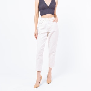 Puede incluir: Una mujer con un top corto de cuello en V de color gris oscuro con un botón dorado y vaqueros de cintura alta de color rosa claro. Lleva tacones altos beiges. El fondo es blanco.