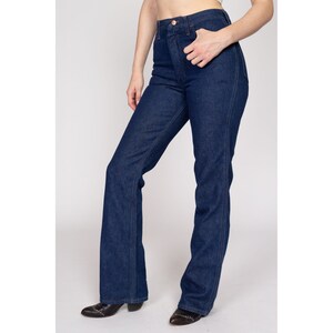 K&ouml;nnte beinhalten: Dunkelblaue Jeans mit hoher Taille und ausgestelltem Bein. Die Jeans haben ein klassisches F&uuml;nf-Taschen-Design und einen Knopf- und Rei&szlig;verschluss. Das Model tr&auml;gt schwarze spitz zulaufende Stiefel.