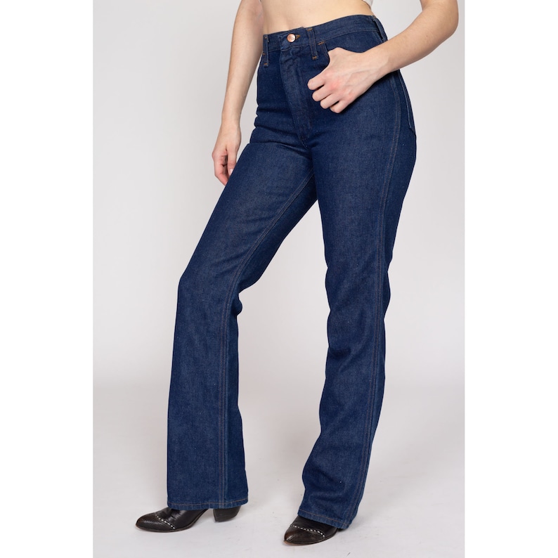 K&ouml;nnte beinhalten: Dunkelblaue Jeans mit hoher Taille und ausgestelltem Bein. Die Jeans haben ein klassisches F&uuml;nf-Taschen-Design und einen Knopf- und Rei&szlig;verschluss. Das Model tr&auml;gt schwarze spitz zulaufende Stiefel.