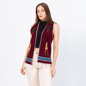 May include: A sleeveless burgundy vest with a Southwestern design. The vest features embroidered cacti and deer, with a white zigzag trim. The bottom has a blue and white striped pattern and a fringe.