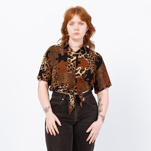 May include: A short-sleeved button-up shirt with a brown, black, and tan leopard print pattern. The shirt is tied at the waist and paired with dark wash jeans. The shirt has a collared neckline.