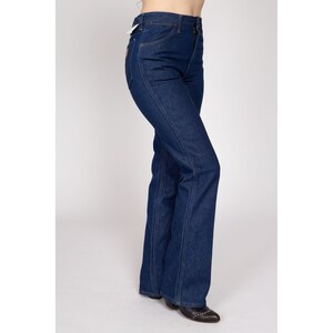 K&ouml;nnte beinhalten: Dunkelblaue Jeans mit hoher Taille und ausgestelltem Bein. Die Jeans haben ein klassisches F&uuml;nf-Taschen-Design mit Rei&szlig;verschluss und Knopfverschluss. Die N&auml;hte sind mit einem kontrastierenden orangefarbenen Faden gen&auml;ht. Die Jeans werden mit einem Etikett gezeigt.