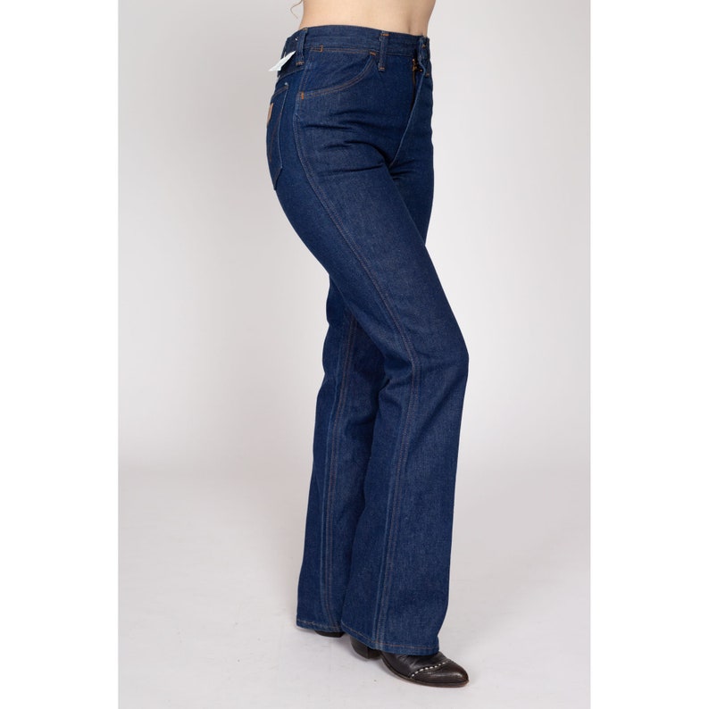 K&ouml;nnte beinhalten: Dunkelblaue Jeans mit hoher Taille und ausgestelltem Bein. Die Jeans haben ein klassisches F&uuml;nf-Taschen-Design mit Rei&szlig;verschluss und Knopfverschluss. Die N&auml;hte sind mit einem kontrastierenden orangefarbenen Faden gen&auml;ht. Die Jeans werden mit einem Etikett gezeigt.
