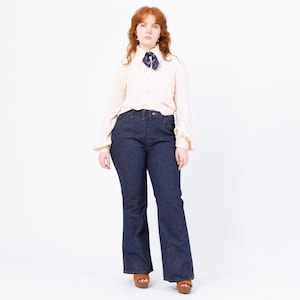 Peut inclure: Une femme porte une chemise crème boutonnée à manches longues et un jean évasé bleu foncé. Un foulard à motifs est noué autour du cou. La femme porte des sandales à plateforme marron.