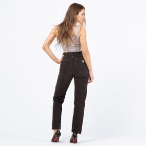 May include: Dark wash denim jeans with a high waist and a small logo on the back pocket. The jeans are paired with a light gray tank top. The model is wearing black high heels.