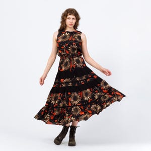 May include: A sleeveless black dress with a floral pattern featuring sunflowers in shades of brown, orange, and yellow. The dress has a tiered skirt and a cinched waist. The model is wearing brown ankle boots.