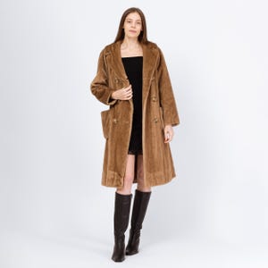 May include: A long, brown faux fur coat with a double-breasted design, large pockets, and button closures. The coat is worn over a black dress and paired with knee-high black boots. The coat's length extends to the mid-calf.