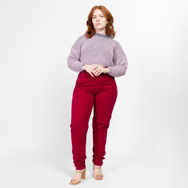 May include: A woman wearing a light purple sweater and high-waisted red jeans. She is standing with her hands clasped in front of her.