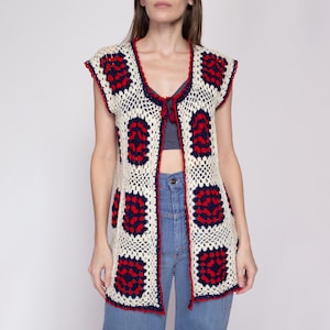 May include: A white crochet vest with red and blue geometric patterns. The vest has a tie closure at the neckline and is sleeveless.