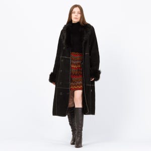 May include: A long black coat with faux fur trim on the collar and cuffs. The coat has large buttons and a tailored fit. The person is wearing a patterned skirt and tall black boots. The coat is a classic outerwear piece.