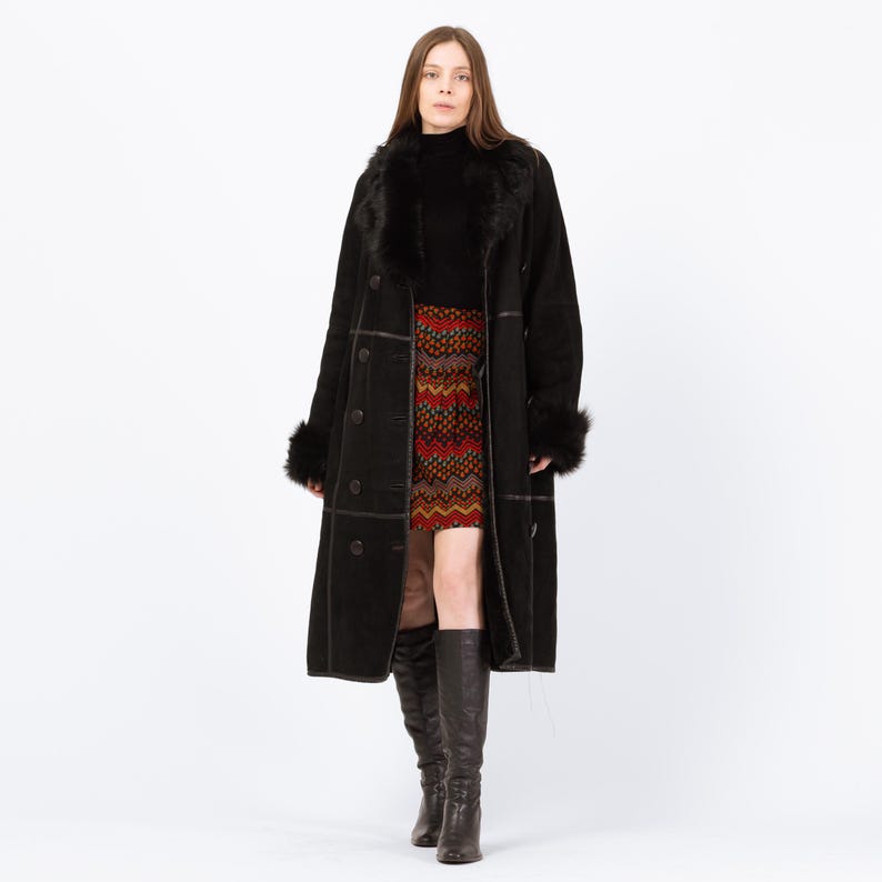 May include: A long black coat with faux fur trim on the collar and cuffs. The coat has large buttons and a tailored fit. The person is wearing a patterned skirt and tall black boots. The coat is a classic outerwear piece.
