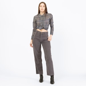 May include: A woman wearing a long-sleeved button-down shirt with a geometric pattern in shades of gray and blue, tied at the waist. She is also wearing gray corduroy pants and black ankle boots. The outfit is set against a white background.