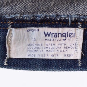 Puede incluir: Primer plano de una etiqueta de vaqueros Wrangler. La etiqueta es blanca con texto negro, incluyendo "Wrangler", "MISSES FULL FIT" e instrucciones de lavado. Los vaqueros son de mezclilla azul oscuro.