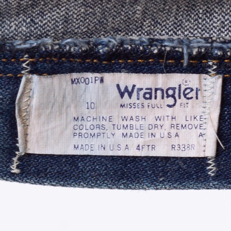 Puede incluir: Primer plano de una etiqueta de vaqueros Wrangler. La etiqueta es blanca con texto negro, incluyendo "Wrangler", "MISSES FULL FIT" e instrucciones de lavado. Los vaqueros son de mezclilla azul oscuro.