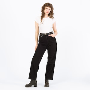 May include: A person wearing a white t-shirt, black jeans, a black belt with a gold buckle, and black boots. The jeans are high-waisted and wide-legged. The person is standing in front of a white background.
