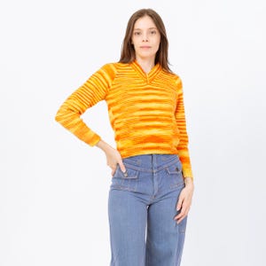 May include: A long-sleeved, orange and yellow striped sweater with a V-neck collar. The sweater is paired with light blue denim jeans with front pockets. The outfit is set against a white background.