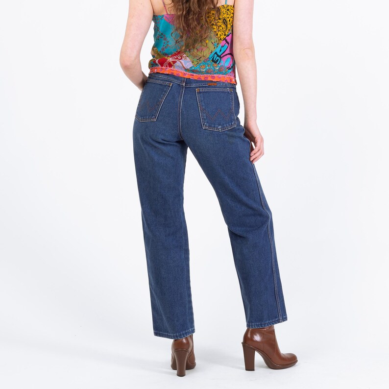 Puede incluir: Vaqueros de mezclilla azul oscuro con corte recto. Los vaqueros tienen un dise&ntilde;o cl&aacute;sico de cinco bolsillos y se combinan con un top estampado colorido y botines marrones con tac&oacute;n. La modelo est&aacute; de espaldas a la c&aacute;mara.