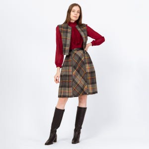 May include: A vintage three-piece skirt suit featuring a plaid pattern in grey, brown, yellow, and red. The outfit comprises a vest, a pleated skirt, and a long-sleeved red blouse. A black belt and tall brown boots complete the ensemble.
