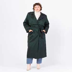 May include: A dark green trench coat with a belted waist and a double-breasted closure. The coat has a classic design with a collar and pockets. The person is wearing blue jeans and light pink boots. The coat is a long length.