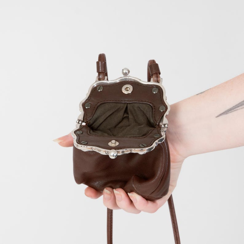 Puede incluir: Un peque&ntilde;o bolso de cuero marr&oacute;n oscuro con un marco de metal plateado y cierre de boquilla. El interior est&aacute; forrado con tela de color oliva oscuro. El bolso cuenta con una correa de cuero fina y un borde festoneado en el marco met&aacute;lico.