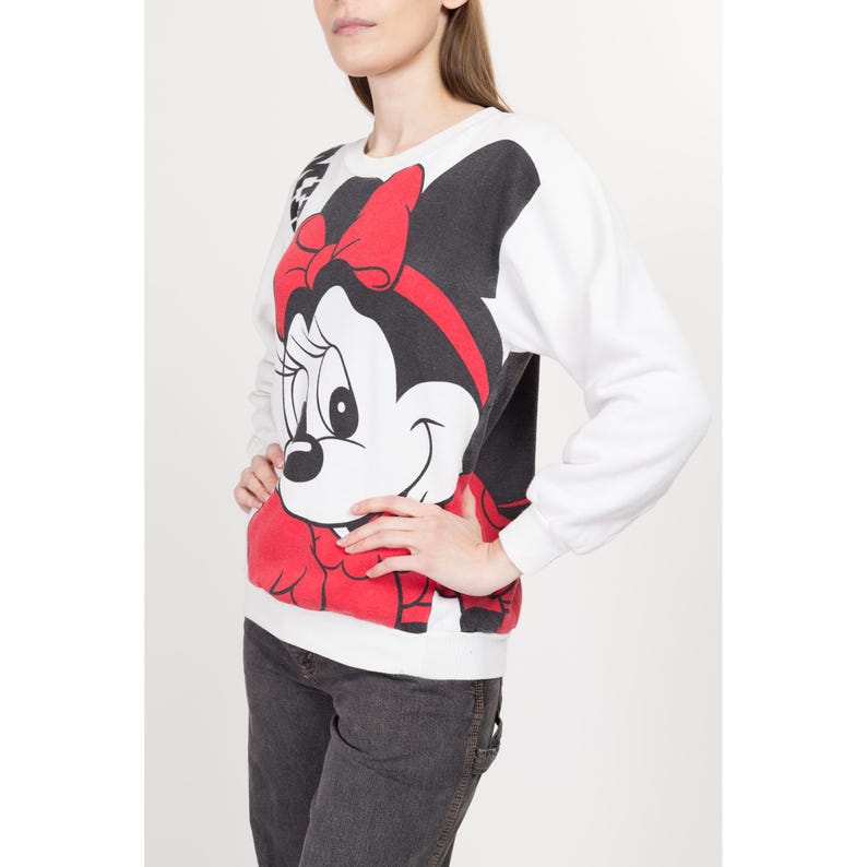 May include: White sweatshirt with a large graphic print of Minnie Mouse. Minnie Mouse is wearing a red bow and has black eyes and a black nose.