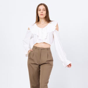 May include: A white, long-sleeved blouse with a ruffled neckline and shoulder cutouts. The blouse has a button-down front and is paired with high-waisted, brown trousers. The trousers have a button and pleat detail.