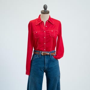 Puede incluir: Una camisa roja de manga larga con botones, costuras blancas en contraste, cuello puntiagudo y dos bolsillos en el pecho, combinada con pantalones vaqueros azules sujetos por un cinturón de cuero marrón con hebilla de metal plateado en un maniquí.