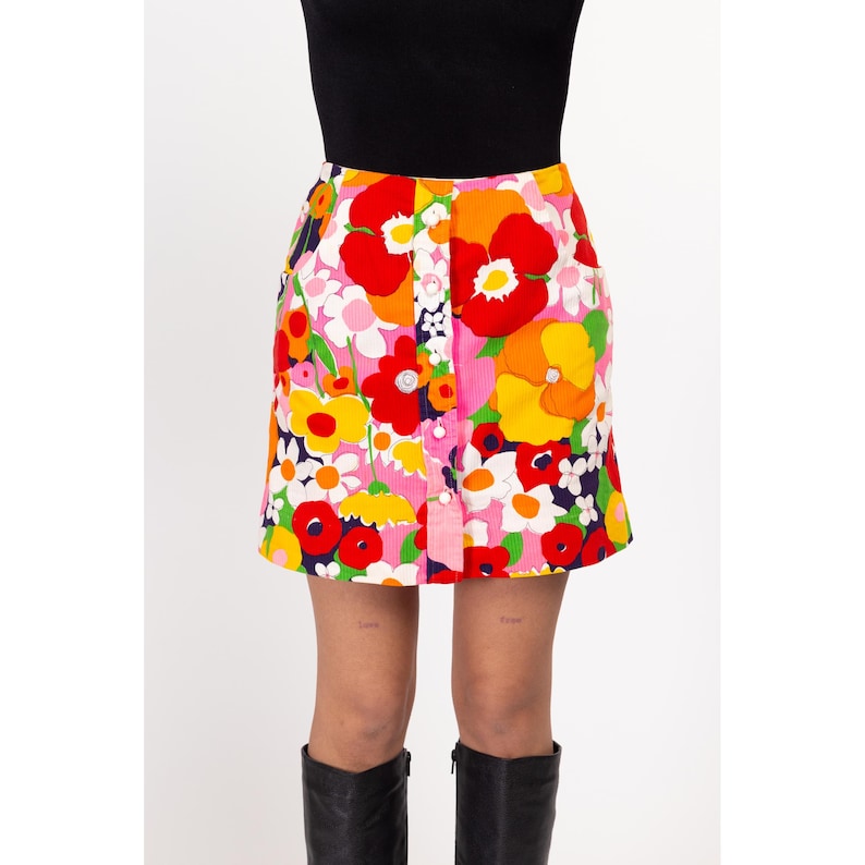 May include: A pink mini skirt with a colorful floral print featuring red, yellow, orange, and white flowers. The skirt has a button closure and a corduroy texture.