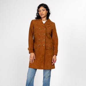 May include: A long brown suede coat with a double breasted closure and gold buttons. The coat has a collar and long sleeves with decorative stitching around the cuffs and pockets.