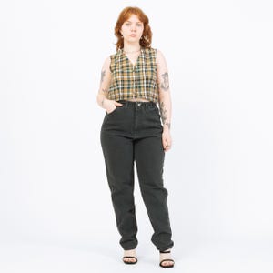 May include: A woman wearing a plaid vest in shades of green, yellow, and brown, paired with dark gray high-waisted jeans. The outfit is completed with black sandals. The overall style is vintage and casual.